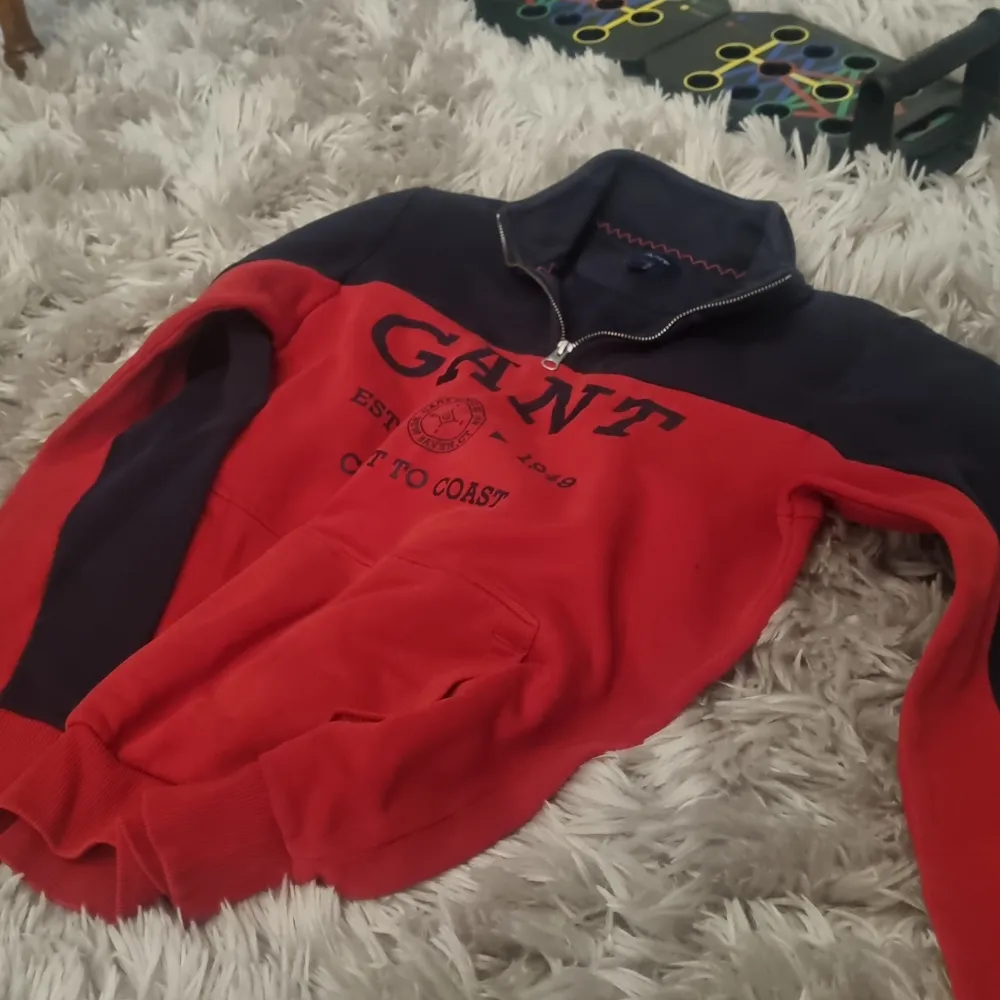Säljer en snygg och bekväm röd och marinblå tröja från GANT. Den har en dragkedja vid halsen och ett tryck med texten 'GANT EST. 1949 COAST TO COAST' på framsidan. Perfekt för en casual look och passar bra till både jeans och chinos. Tröjan är i bra skick och har anvädns sällan. Hupparit & Collegepaidat.