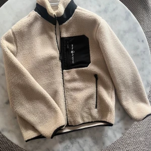 Beige fleecejacka från Sail Racing - Säljer en supermysig beige fleecejacka från Sailracing. Den har en dragkedja framtill och en cool svart bröstficka med sailrace-loggan. Perfekt för kyliga dagar och har en skön passform med långa ärmar. Inga defekter✅