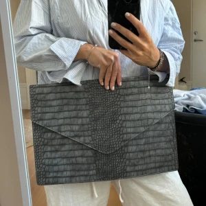 Grå kuvertväska från Bianca - Säljer en stilren grå kuvertväska från Bianca med ett snyggt krokodil-mönster. Väskan har en elegant design och är perfekt för att bära dokument eller en dator. Den har en diskret stängning och en liten metallplatta med märkesloggan på framsidan.