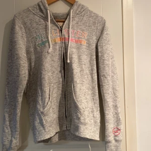 Grå hoodie från Hollister - Grå kofta från Hollister med färgglatt tryck på bröstet. Den har en dragkedja och justerbar huva med snören.  Bra skick. 