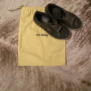 Svarta sneakers från Axel Arigato - Säljer ett par svarta sneakers från Axel Arigato med fågelbroderi på sidan. Skorna har snörning och en vit sula. Perfekta för en avslappnad stil.