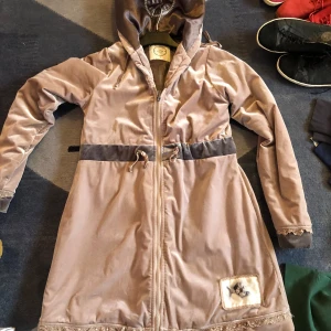 Beige parkajacka med huva - Snygg beige parkajacka med dragkedja och huva. Jackan har en justerbar midja med snörning och en dekorativ patch längst ner. Perfekt för kyligare dagar.