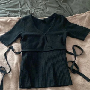 Svart ribbad topp med knytband - Snygg svart topp med v-ringning och trekvartsärm. Den har en ribbad nederdel och knytband i midjan för en stilren look. Perfekt för både vardag och fest.