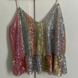 Glittrig regnbågsrandig topp från Zara - Säljer en glittrig topp från Zara med regnbågsrandigt mönster. Toppen har smala axelband och en lätt volangdetalj vid nederkanten. Perfekt för fest eller konserter. Säkert superfin till dom kommande Sabrina Carpenter konserterna 💋