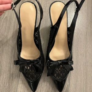 Svarta klackskor med rosett - Eleganta svarta klackskor med spetsig tå och dekorativ rosett. Skorna har en stilren design med spetsdetaljer och en justerbar rem runt hälen. Perfekta för en chic look.