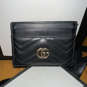 Svart kortfodral från Gucci - Snyggt svart kortfodral från Gucci i skinn med quiltad design och det ikoniska GG-emblemet i guld. Perfekt för att hålla dina kort organiserade med stil. Orginalpris 3300kr säljs för 2000kr. Kartong + påse medföljer.