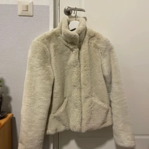 Beige teddyjacka från ONLY - Mysig beige teddyjacka från ONLY med hög krage och knappar. Perfekt för kyliga dagar med sin mjuka och varma design. Jackan har två praktiska fickor framtill.