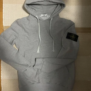 Grå hoodie från Stone Island - Snygg grå hoodie från Stone Island med klassisk logga på ärmen. Perfekt för en avslappnad stil. Mjuk och bekväm med justerbar huva och känguruficka framtill. Priset är 949kr hör av er vid frågor👏😁 Storlek M, varan är i nyskick och har knappt används, i princip nytt. 
