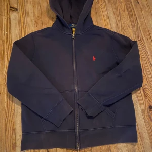 Marinblå hoodie från Ralph Lauren - Säljer en stilren marinblå hoodie från Ralph Lauren med dragkedja och det klassiska röda broderade logotypen på bröstet. ‼️Lappen med storlek har blivit av klippt och tröjan har blivit lite dissfärgad av tvättningar‼️ där utav det billiga priset. Tröjan är i perfekt kondition utom lappen då. Storlek är size M 10-12 i barnstorlek