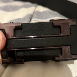 Svart bälte med H metallspänne - Snyggt svart bälte med ett robust metallspänne. Bältet har en stilren design med en diskret logotyp präglad på ytan. Perfekt för att ge en elegant touch till din outfit.