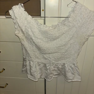 Vit topp från Gina Tricot - Säljer en söt vit topp från Gina Tricot i storlek XS. Toppen har en smockad design med volangdetaljer vid ärmslut och nederkant. Perfekt för en somrig look!