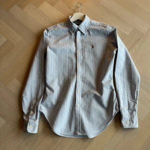 Randig oxfordskjorta från Ralph Lauren - Snygg och stilren randig skjorta från Ralph Lauren i skinny fit. Skjortan är tillverkad i 100% bomull och har en klassisk krage med knappar. Perfekt för en elegant look. Ena knappen vid kragen saknas, lätt gul i ärmhålor. Nypris 1800 kr