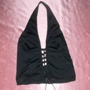 Svart halterneck topp med snörning - Snygg svart halterneck topp med snörning framtill. Perfekt för en kväll ute eller en fest. Toppen har en djup urringning och är tillverkad i ett mjukt material för en bekväm passform.