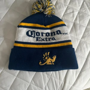 Blå och gul mössa med Corona Extra-logga - Snygg mössa i blått och gult med Corona Extra-logga. Den har en stor tofs på toppen och ett broderat lejonmotiv framtill. Perfekt för kalla dagar.