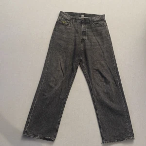 Grå baggy jeans från J&J - Säljer ett par grå jeans med baggy passform. De har en klassisk femficksdesign och en knappgylf. Perfekta för en avslappnad stil. Bra skick men lite slitna vid byxöppningen längst ner på ena benet