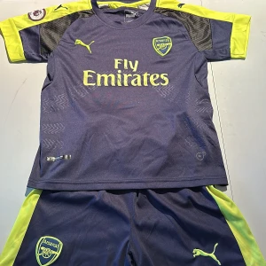 Arsenal fotbollströja med shorts - Säljer en Arsenal fotbollströja med korta ärmar och matchande shorts. Tröjan är mörkblå med gula detaljer och har 'Fly Emirates' tryckt på framsidan. Baksidan har namnet 'Alexis' och nummer 7. Trycket på framsidan är slitet, se bild. Perfekt för fans av Arsenal! 💛