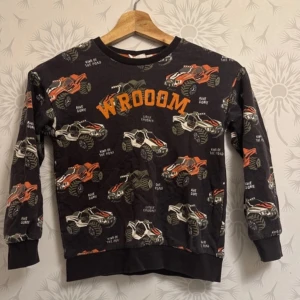 Svart tröja med biltryck - Cool svart tröja med färgglatt biltryck och texten 'WROOM' i orange. Perfekt för bilentusiaster! Tröjan har långa ärmar och rund halsringning.  Tröjan är hel de finns uterst en liten  hål i armen som har blivet sytt.