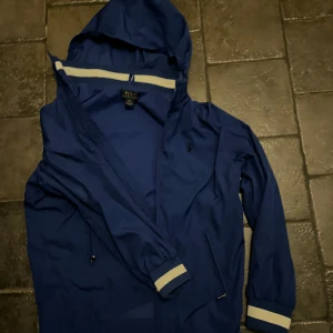 Blå vindjacka från Polo Ralph Lauren - Snygg blå vindjacka från Polo Ralph Lauren med huva och dragkedja. Jackan har vita detaljer vid ärmslut och huva, samt två fickor med dragkedja. Perfekt för blåsiga dagar.