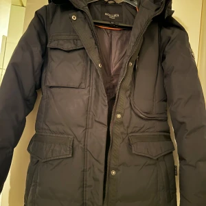 Svart parkajacka från Hollies - Säljer en svart parkajacka från Hollies med flera praktiska fickor och dragkedja. Jackan har långa ärmar och är perfekt för kyligare väder. Den har en stilren design och är både funktionell och snygg. Använd endast 1 vinter