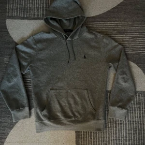Grå hoodie från Ralph Lauren - Säljer en stilren grå hoodie från Ralph Lauren med klassisk logga på bröstet. Tröjan har en bekväm passform med långa ärmar och en praktisk magficka. Perfekt för en avslappnad look.