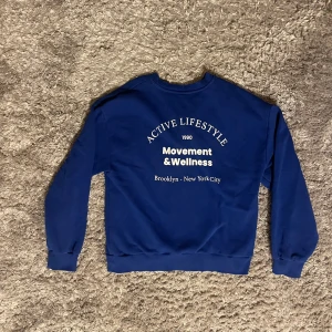 Blå sweatshirt från Gina Tricot - Säljer en blå sweatshirt från Gina Tricot med texten 'Active Lifestyle Movement & Wellness Brooklyn - New York City' tryckt på både fram- och baksidan. Tröjan har långa ärmar och en rund halsringning. Perfekt för en avslappnad stil.