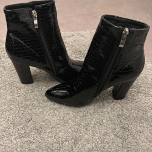 Svarta stövletter med dragkedja - Snygga svarta stövletter med krokodilmönster och dragkedja på sidan. De har en spetsig tå och en stabil klack som ger en elegant look. Perfekta för att lyfta vilken outfit som helst.