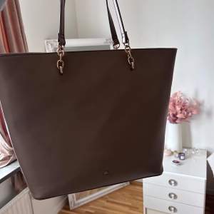 Snygg brun axelväska i skinn med stilrena detaljer. Väskan har två långa axelremmar och små metallbeslag som ger en elegant touch. Perfekt för att bära med sig allt man behöver under dagen.