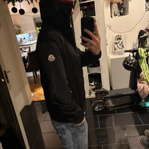 Svart jacka från Moncler - Säljer en stilren svart jacka från Moncler med huva och dragkedja. Jackan har en broderad logotyp på ärmen och en röd-vit randdetalj på huvan. Perfekt för kyligare dagar. Nfc Scan och äktehetsbevis finns!! Cond är 9/10 då jag bara använt den under hösten men är sugen på något annat. För mer frågor skriv gärna till mig/mvh Simon 