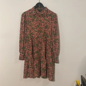 Blommig klänning från Monki - Charmig blommig klänning från Monki i storlek XS. Klänningen har ett färgglatt mönster med rosa, gröna och bruna blommor. Den har långa ärmar och knappar framtill, vilket ger en söt och avslappnad stil.