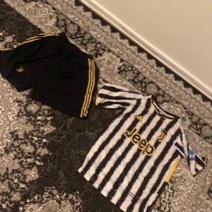 Juventus fotbollströja med shorts - Säljer en Juventus fotbollströja med Pogba 10 på ryggen. Tröjan är randig i svart och vit med gula detaljer och korta ärmar. Matchande svarta shorts med gula ränder ingår.