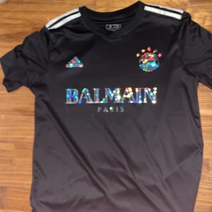 Svart fotbollströja med Balmain-logga - Snygg svart fotbollströja från Adidas med Balmain Paris-logga i holografiskt tryck. Tröjan har korta ärmar och klassiska tre ränder på axlarna. Perfekt för dig som vill sticka ut på planen! Bayern muchen är laget❤️🤍💙 kan gå ner i pris😘