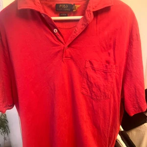 Polo Ralph Lauren pike  - Snygg röd piké från Polo Ralph Lauren med korta ärmar och en bröstficka. Perfekt för en avslappnad stil. Tillverkad i mjukt material för extra komfort.