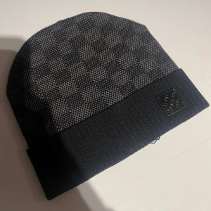 mössa - Snygg svart och grå mössa med rutigt mönster. Den har en bred ribbad kant och en diskret logotyp framtill. Perfekt för kyliga dagar.