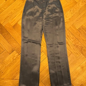 Grå byxor från Ahlvar Gallery - Snygga grå byxor från Ahlvar Gallery i ett glansigt material. De har en hög midja och raka ben, vilket ger en elegant look. Byxorna är försedda med en knapp och dragkedja framtill.