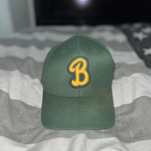 Grön keps med gul bokstav - Snygg grön keps med en broderad gul bokstav 'B' på framsidan. Perfekt för en avslappnad stil. NBA lag keps.