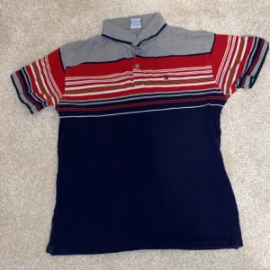 Randig pikétröja Tommy Hilfiger - Snygg pikétröja från Tommy Hilfiger med ränder i rött, vitt och blått. Tröjan har en klassisk krage och korta ärmar, perfekt för en avslappnad stil.