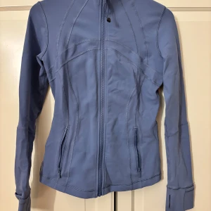 Blå jacka från Lululemon - Snygg blå jacka från Lululemon med dragkedja framtill och figurnära passform. Perfekt för träning eller vardagsbruk. Jackan har långa ärmar och praktiska fickor med dragkedja. Köpt av en annan tjej som köpte den i new York för 2200 kr och endast använd 1 gång. Obs! Färgen går ej att köpa mer heller ☺️