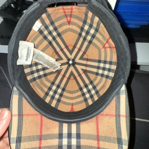 Svart keps från Burberry - Snygg svart keps från Burberry med broderad logotyp på framsidan. Insidan har det klassiska Burberry-rutiga mönstret. Perfekt för en stilren look. Storlek S