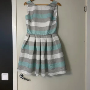 Randig ärmlös klänning - Snygg ärmlös klänning med breda ränder i grått, vitt och ljusblått. Klänningen har en rund hals och en v-formad rygg med dragkedja. Perfekt för en somrig look!