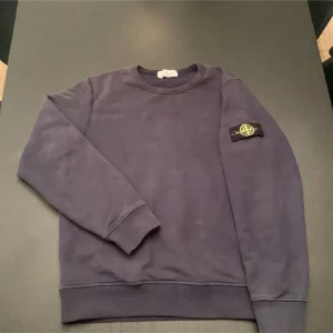 Mörkblå sweatshirt från Stone Island Junior - Säljer en stilren mörkblå sweatshirt från Stone Island Junior. Tröjan har en rund halsringning och ett ikoniskt märke på ärmen. Perfekt för en casual look.