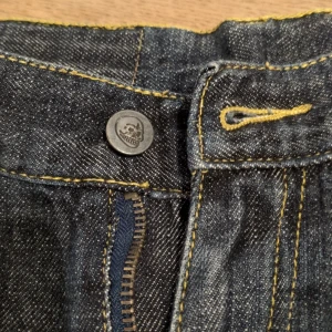 Jeansshorts från Cheap Monday - Snygga jeansshorts från Cheap Monday med råa kanter och cool dödskalleknapp. De har en klassisk femficksdesign och en mörk tvätt med kontrasterande sömmar. Perfekta för en avslappnad stil.