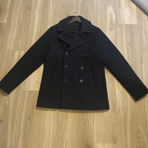 Marin blå skepparkavaj - Snygg marinblå skepparkavaj med dubbelknäppning och klassisk krage. Perfekt för kyligare dagar med sin stilrena design och långa ärmar. Passar både till vardags och mer uppklädda tillfällen.