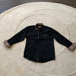 Svart skjorta från Burberry - Snygg svart skjorta från Burberry med klassisk krage och långa ärmar. Den har stiliga rutiga detaljer på insidan av kragen och ärmsluten. Perfekt för en elegant look.     Skjortans storlek är 164 men passar S. Fråga gärna om mer bilder