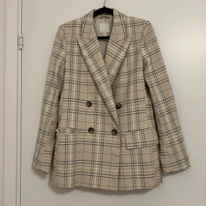 Rutig kavaj från H&M - Snygg rutig kavaj från H&M i beige och svart. Dubbelknäppt design med klassisk krage och långa ärmar. Perfekt för en stilren look. Ordentligt och robust material. Oversize, passar 34-36