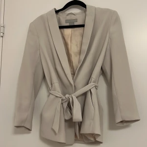 Beige kavaj från H&M - Elegant beige/vit kavaj från H&M med knytband i midjan. Kavajen har en stilren design med långa ärmar och en mjuk, bekväm passform. Perfekt för en chic och sofistikerad look.