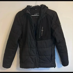Svart jacka från Sail Racing - Snygg svart jacka från Sail Racing med dragkedja och huva. Perfekt för kyligare dagar med sin quiltade design. Jackan har en praktisk bröstficka med dragkedja och är tillverkad i slitstarkt material.pris går att diskutera