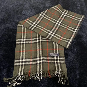 Rutig halsduk från Burberry - Snygg rutig halsduk från Burberry i klassiska färger. Den har en bas i olivgrön med inslag av rött, svart och vitt. Perfekt accessoar för att hålla sig varm och stilren under kyliga dagar. 100% cashmere
