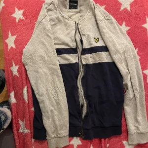Grå och marinblå tröja från Lyle & Scott - Säljer en snygg grå och marinblå tröja från Lyle & Scott med dragkedja och broderad logga på bröstet. Tröjan har långa ärmar och ribbade muddar. Perfekt för en casual look. Har lite mini slitage. Väl använt. 