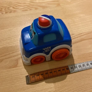 Leksaksbil - Blå leksaksbil med stora oranga hjul och ett glatt ansikte framtill. Bilen har en röd och blå siren på taket. När man ger den lite fart kan den köra själv en bit samt att sirenen då snurrar.