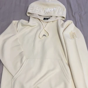 Beige hoodie från Mackage - Säljer en stilren beige hoodie från Mackage med broderad logga på huvan. Tröjan har en klassisk känguruficka och justerbar dragsko i huvan. Perfekt för en avslappnad stil.